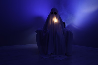 Creepy ghost. Woman covered with sheet sitting in armchair in blue light