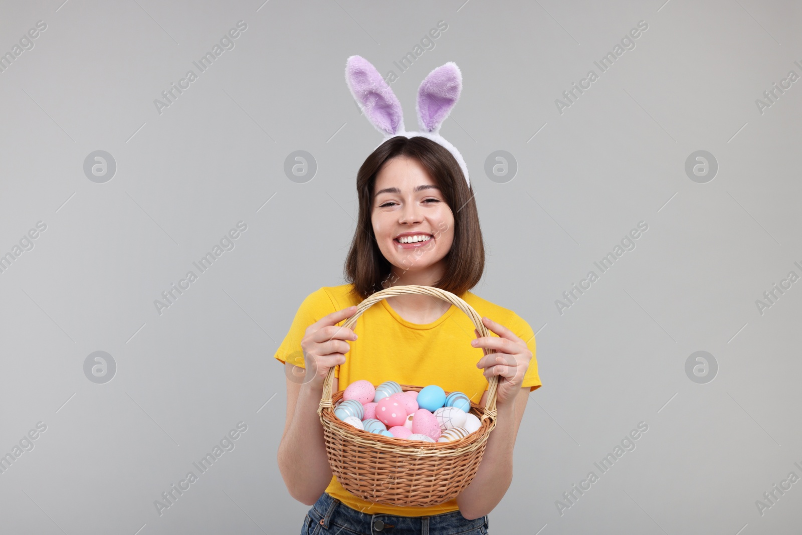 Photo of Easter celebration. Happy woman with bunny ears and wicker basket full of painted eggs on grey background