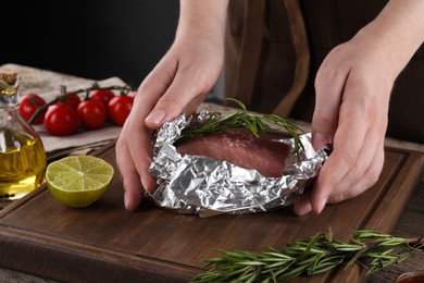 Woman wrapping meat with rosemary in aluminum foil at wooden table, closeup