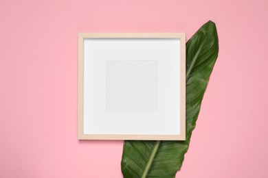 Photo of Empty photo frame and green leaf on pink background, flat lay. Mockup for design