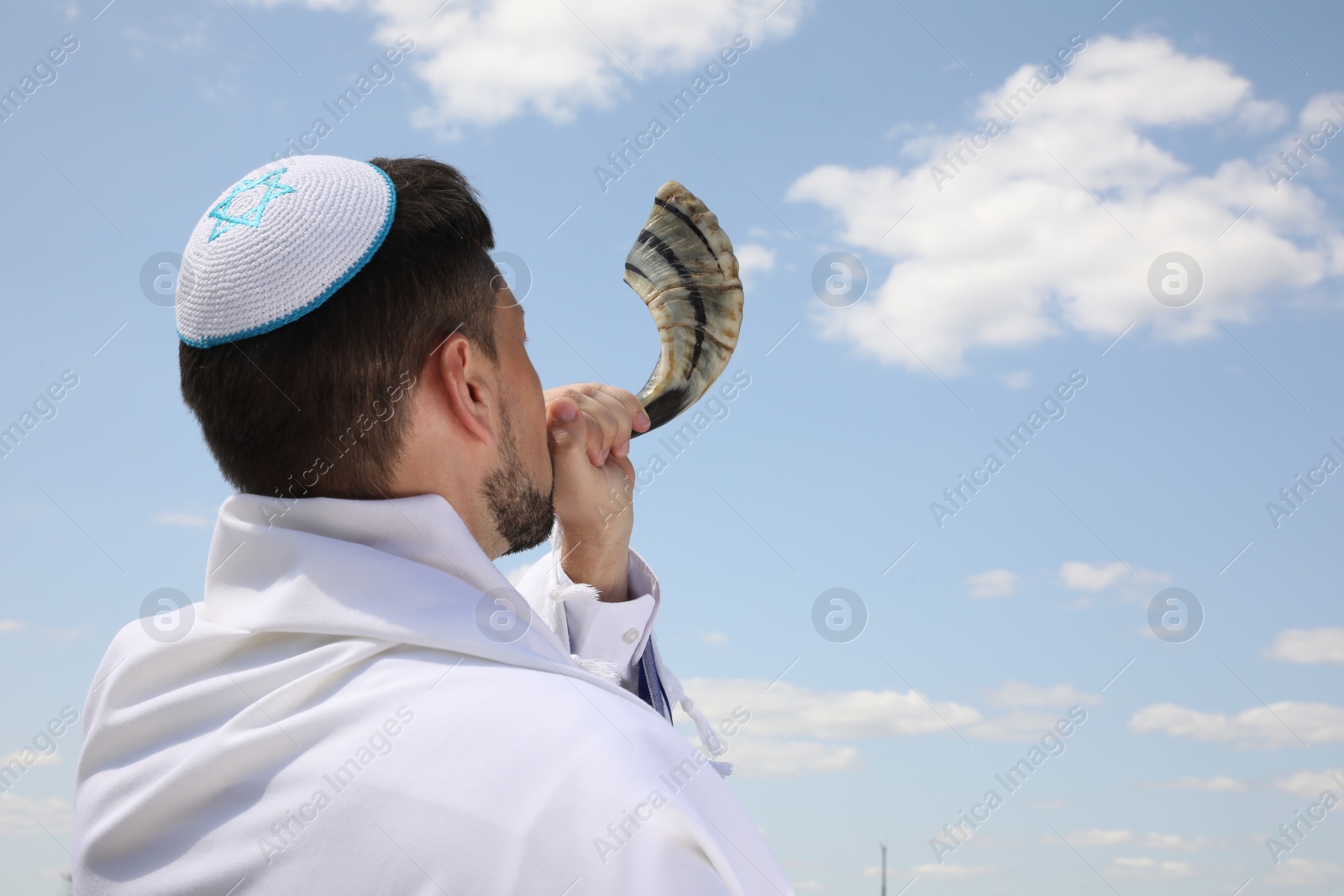 Photo of Jewish man in kippah and tallit blowing shofar outdoors. Rosh Hashanah celebration