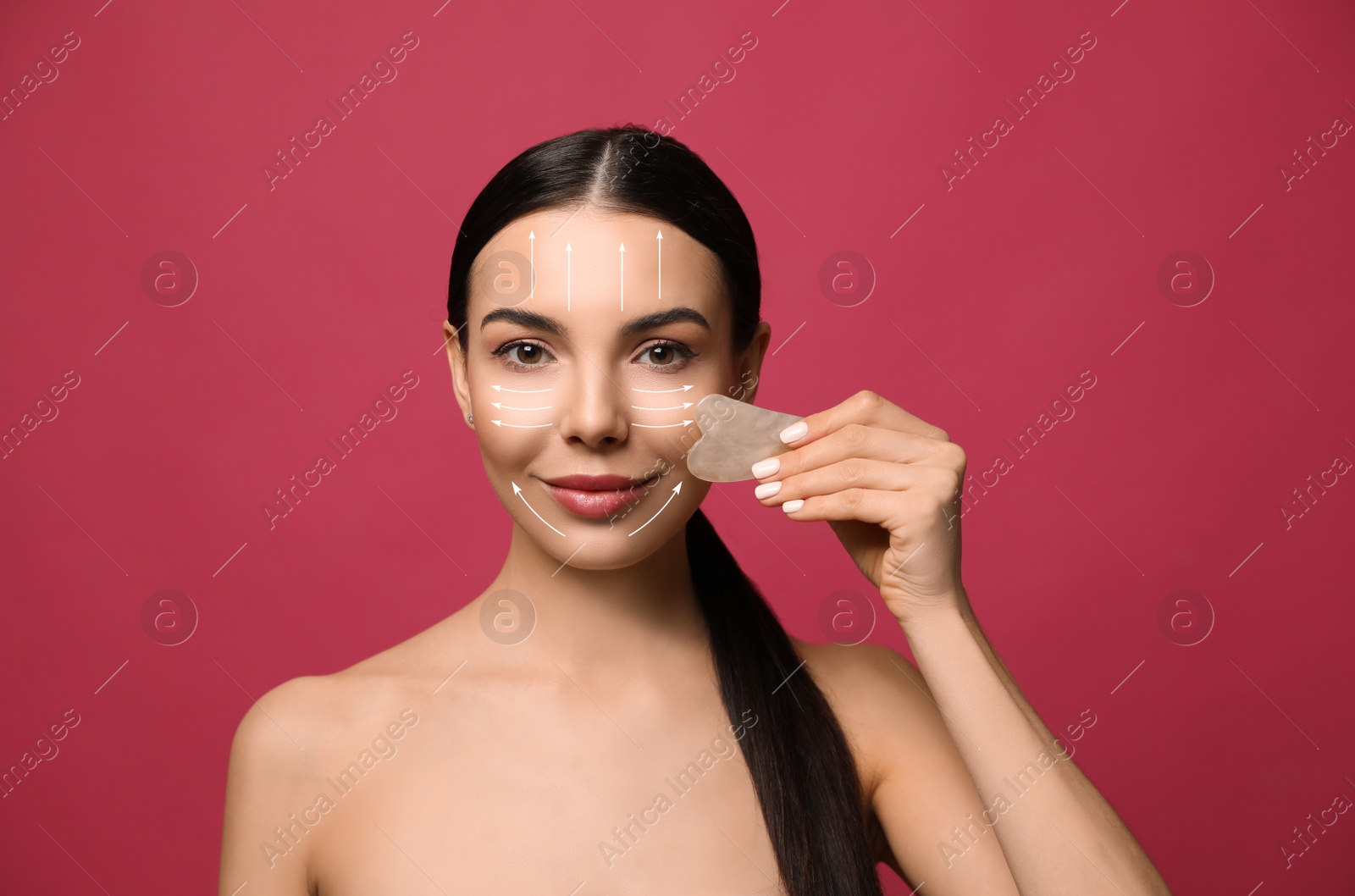 Image of Beautiful young woman doing facial massage with gua sha tool on crimson background