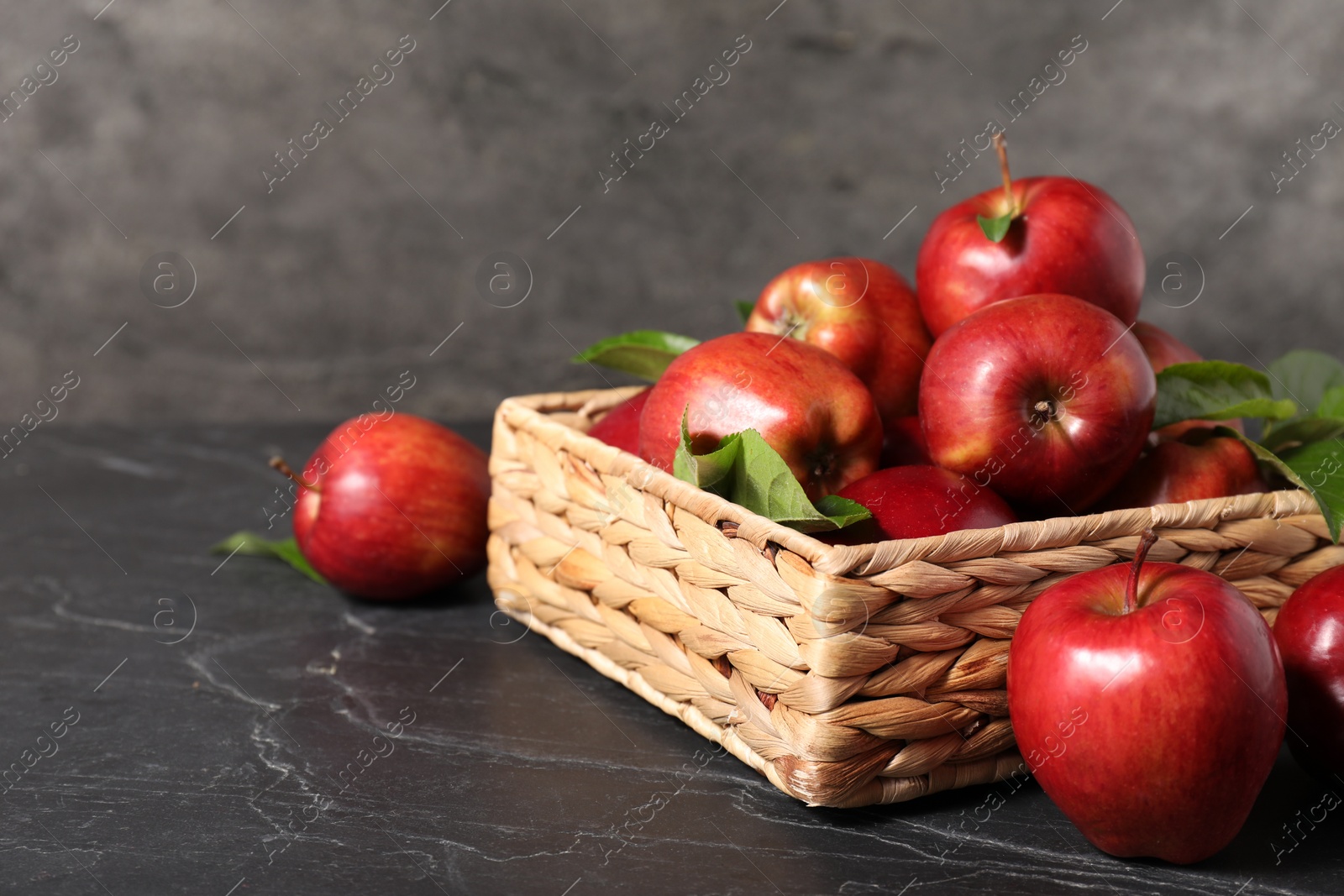 Photo of Fresh red apples and leaves in basket on dark grey table. Space for text