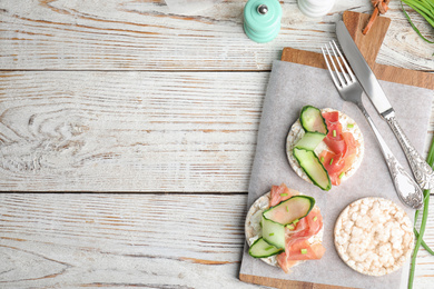 Photo of Puffed rice cakes with prosciutto and cucumber served on white wooden table, flat lay. Space for text
