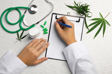 Female doctor with clipboard, hemp leaves and stethoscope at white wooden table, top view