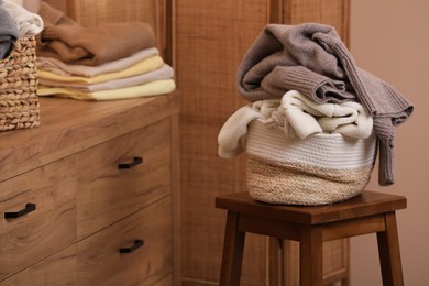 Wicker laundry basket overfilled with clothes on wooden stool indoors