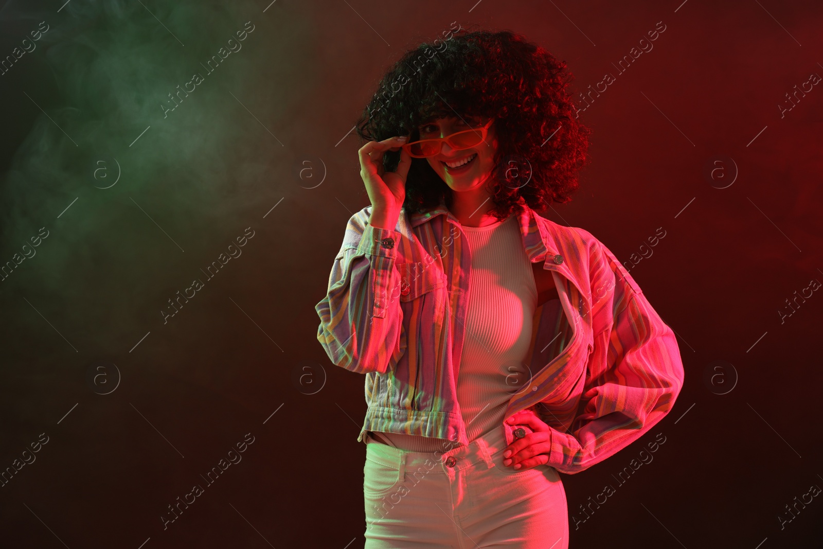 Photo of Beautiful young woman in sunglasses posing on color background in neon lights and smoke
