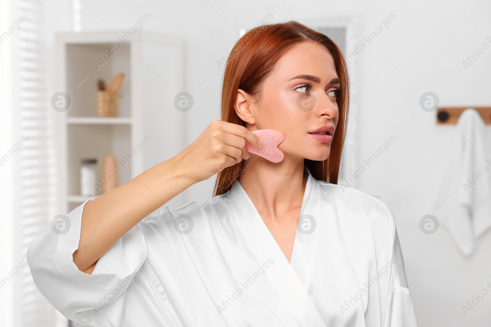 Photo of Young woman massaging her face with rose quartz gua sha tool in bathroom