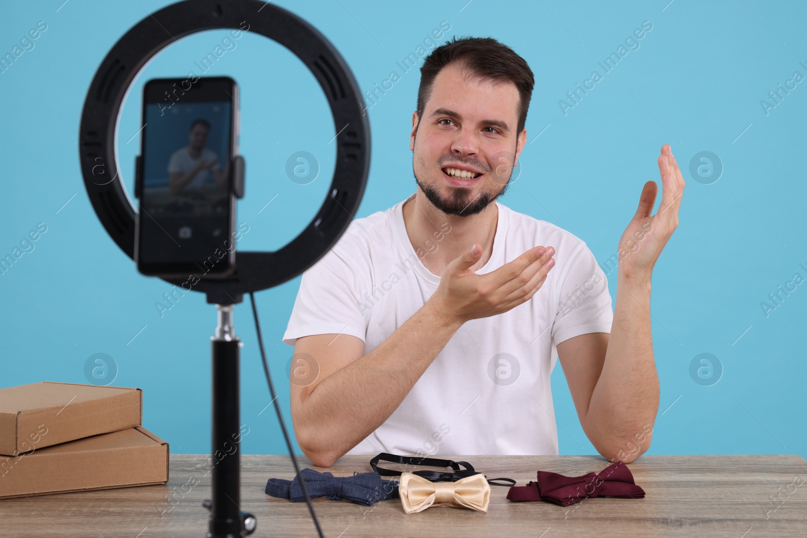 Photo of Smiling fashion blogger showing bow ties while recording video at table against light blue background