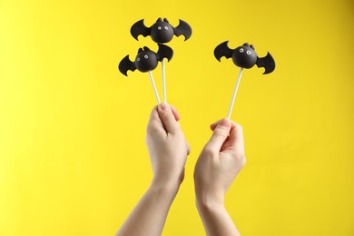 Photo of Woman with delicious bat shaped cake pops on yellow background, closeup. Halloween celebration