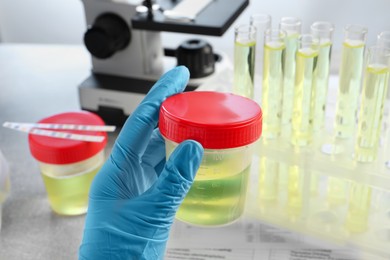 Doctor holding container with urine sample for analysis at grey table in laboratory, closeup