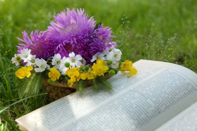 Photo of Bouquet of beautiful wildflowers and open book on green grass outdoors, closeup