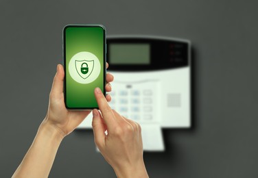 Woman operating home alarm system via mobile phone against dark wall with security control panel, closeup. Application with illustration of padlock in shield on device screen