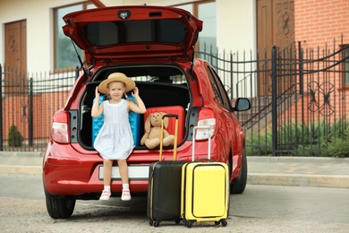 Little girl sitting in car trunk with suitcases outdoors. Space for text
