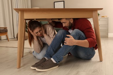 Photo of Scared couple hiding under table in living room during earthquake