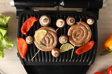 Photo of Electric grill with homemade sausages and vegetables on wooden table, flat lay