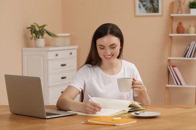 Young woman holding cup with coffee and writing in notebook at wooden table indoors