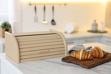 Photo of Wooden bread box and board with croissants on white marble table in kitchen