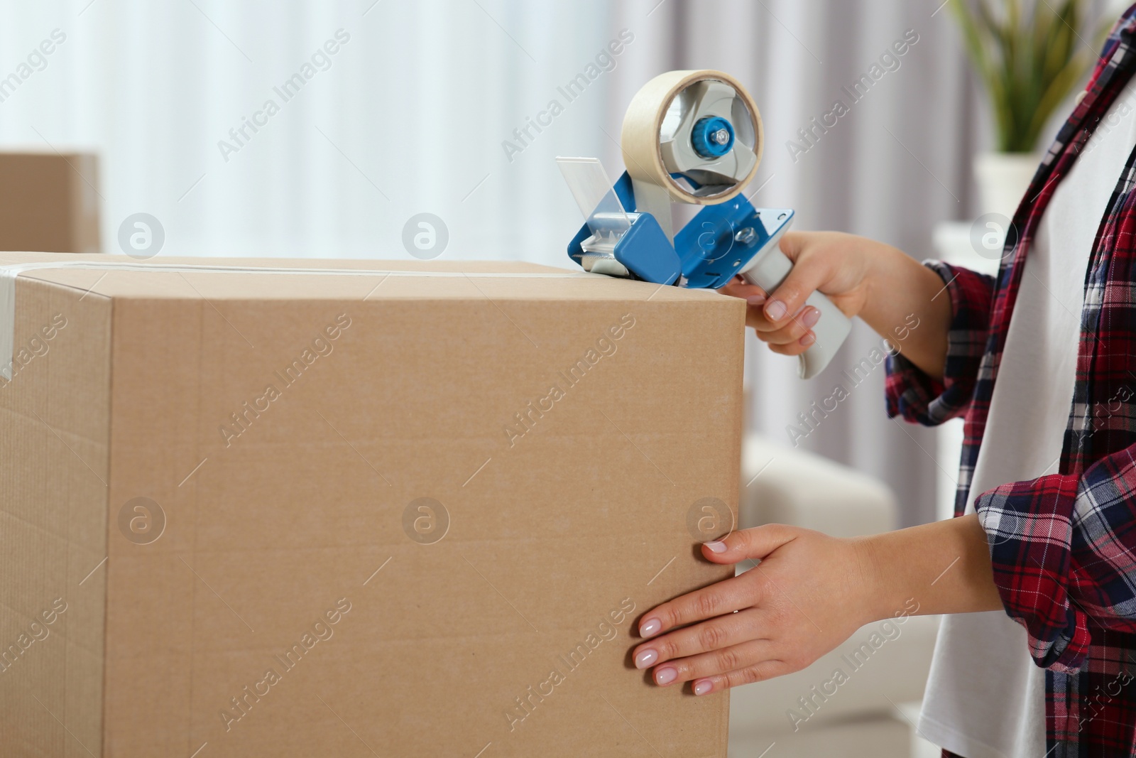 Photo of Woman applying adhesive tape on box with dispenser indoors, closeup