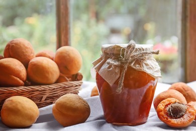 Photo of Jar of delicious jam and fresh ripe apricots on tablecloth indoors. Fruit preserve