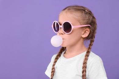 Photo of Girl in sunglasses blowing bubble gum on purple background, space for text
