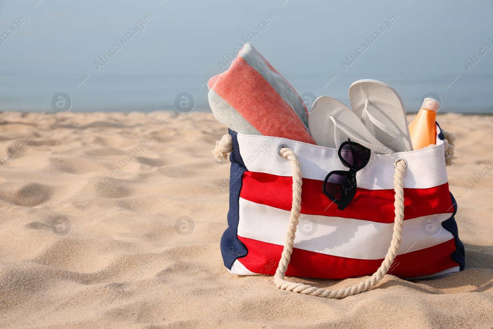 Photo of Beach bag with flip flops, towel, sunglasses and sunscreen on sandy seashore, space for text