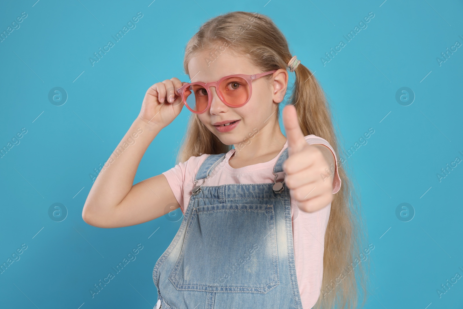 Photo of Girl in pink sunglasses showing thumb up on light blue background