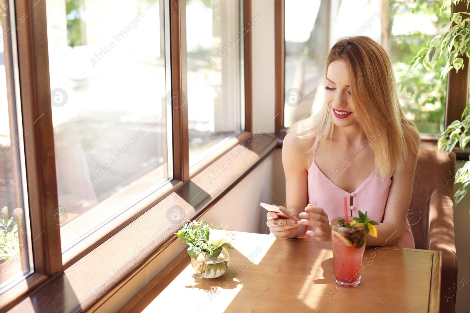 Photo of Young woman using mobile phone while drinking tasty natural lemonade at table in cafe. Detox drink