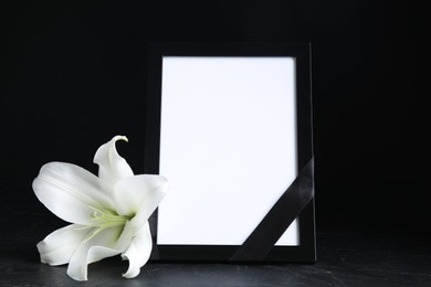 Funeral photo frame with ribbon and white lily on black table against dark background. Space for design