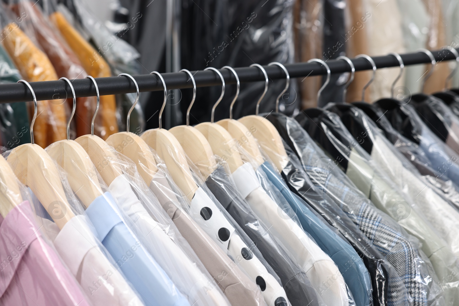 Photo of Dry-cleaning service. Many different clothes in plastic bags hanging on rack indoors, closeup