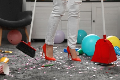 Photo of Woman with sweep broom and dust pan cleaning messy room after party, closeup view of legs