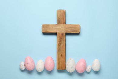 Wooden cross and painted Easter eggs on light blue background, flat lay