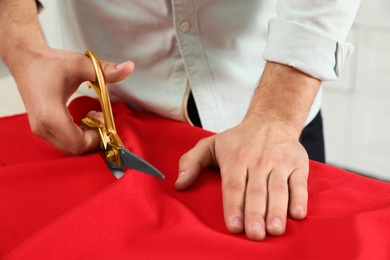 Photo of Professional tailor cutting red fabric with scissors in workshop, closeup