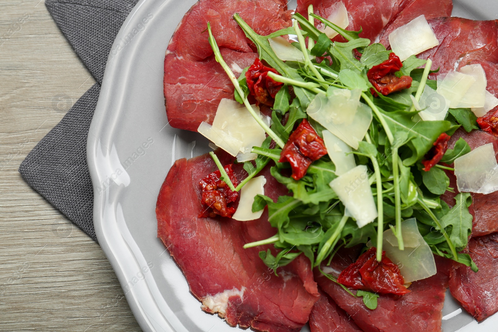 Photo of Plate of tasty bresaola salad with sun-dried tomatoes and parmesan cheese on wooden table, top view