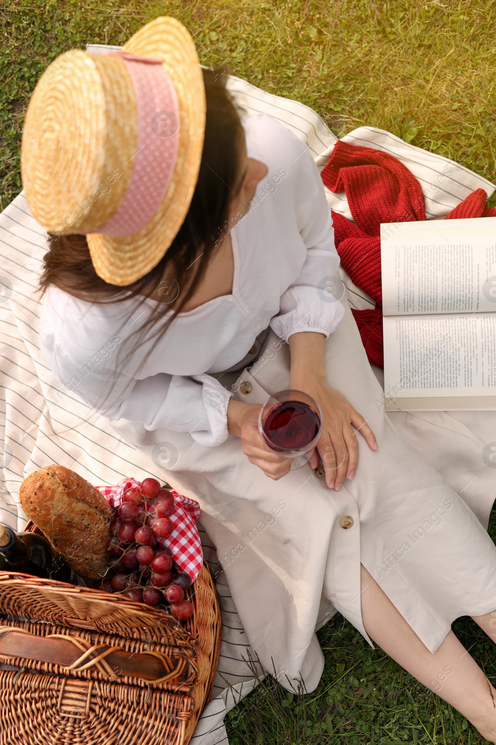 Photo of Woman with glass of wine, book and picnic basket on green grass, above view