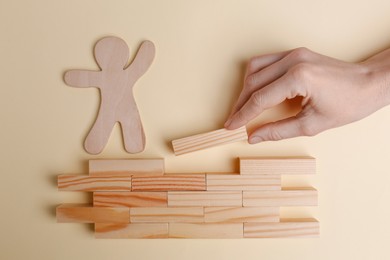 Woman putting wooden block to help human figure cross construction on beige background, top view