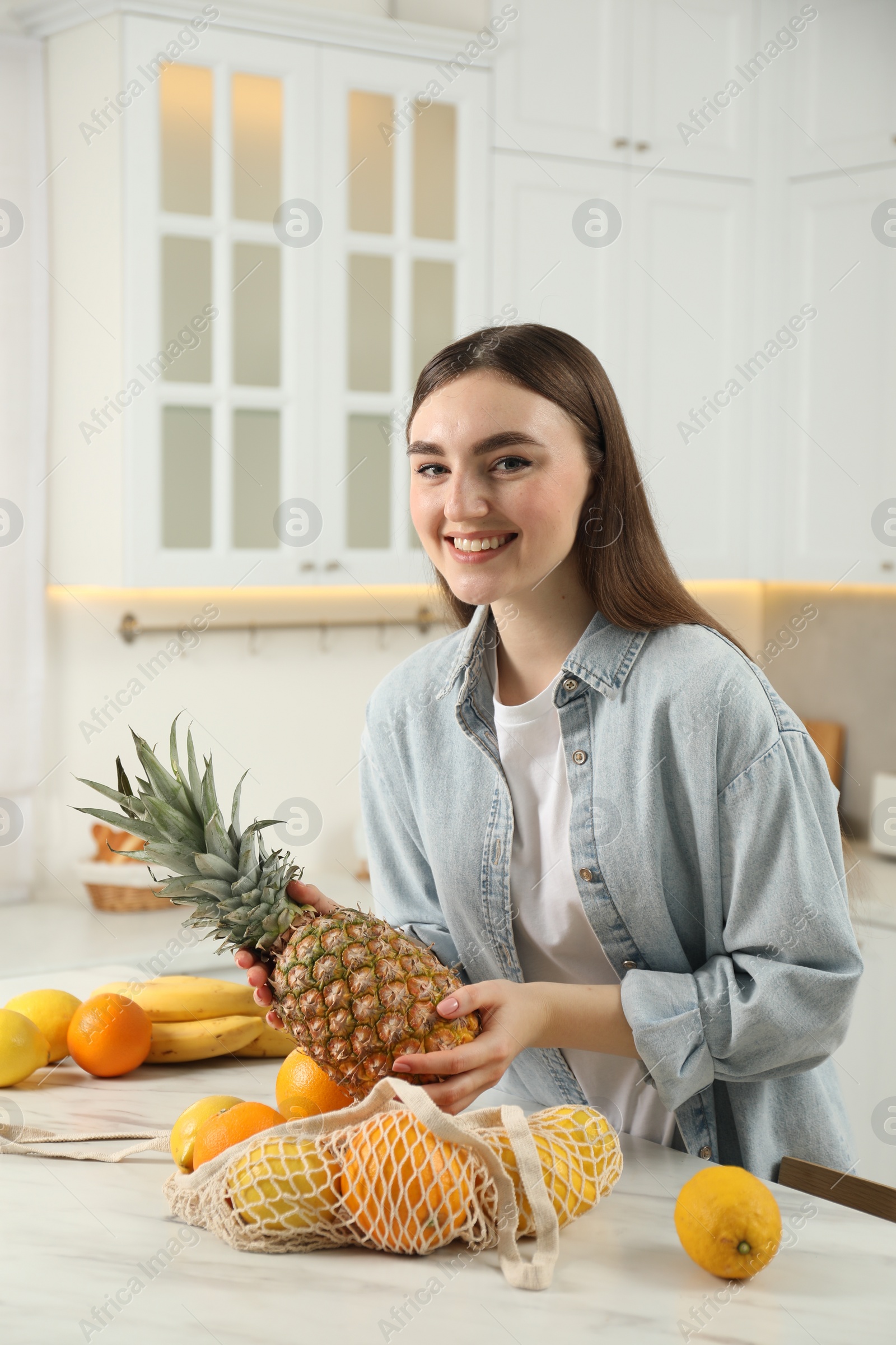 Photo of Woman taking pineapple out from string bag at light marble table in kitchen