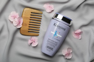 Photo of MYKOLAIV, UKRAINE - SEPTEMBER 07, 2021: Kerastase shampoo, hair comb and hydrangea florets on grey fabric, flat lay