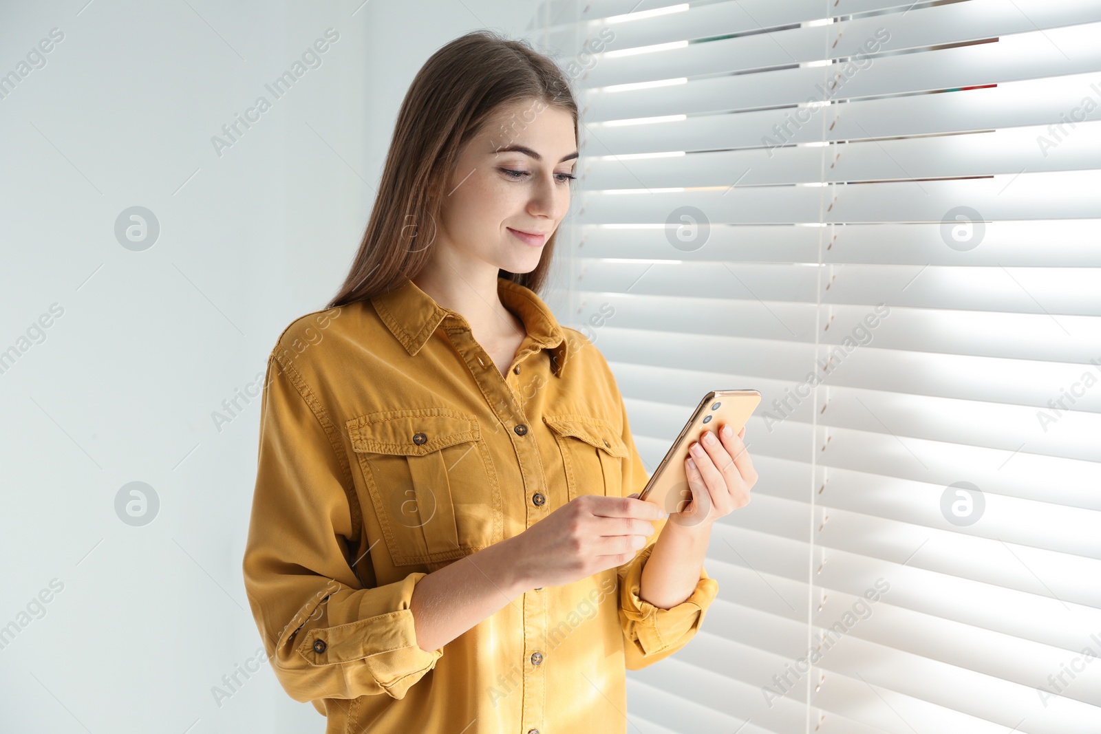 Photo of Young woman using modern smartphone near window at home