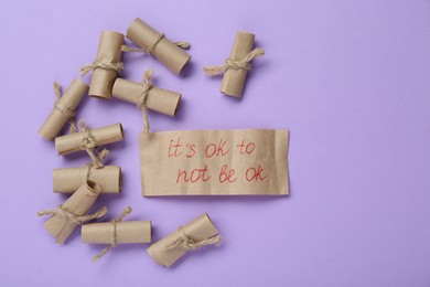 Card with phrase It`s Ok to Not Be Ok and rolled pieces of paper on violet background, flat lay. Space for text