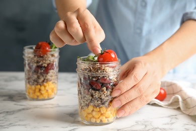 Photo of Woman putting tomato into jar with healthy quinoa salad and vegetables on table, closeup