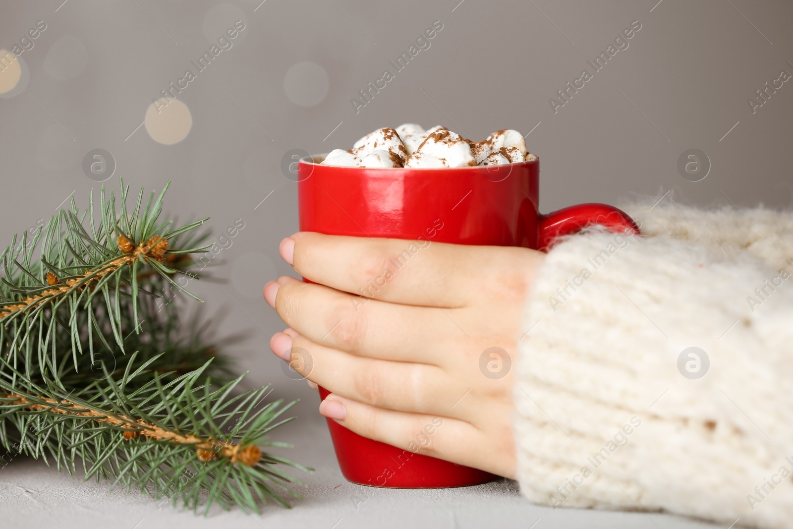 Photo of Woman holding cup of tasty cocoa with marshmallows at white table against blurred lights, closeup