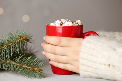 Woman holding cup of tasty cocoa with marshmallows at white table against blurred lights, closeup