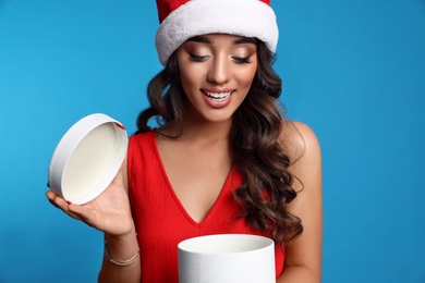Photo of Beautiful woman in Santa hat opening Christmas gift on blue background