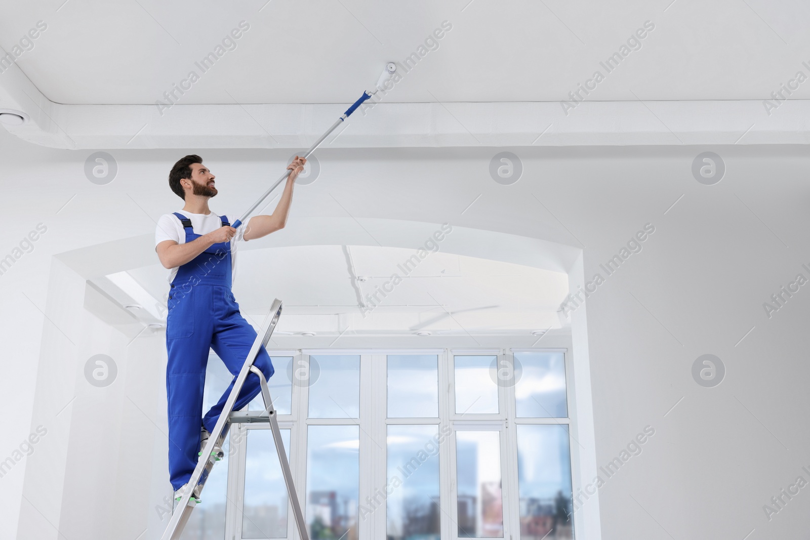 Photo of Handyman painting ceiling with roller on step ladder in room