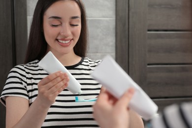 Young woman squeezing toothpaste from tube onto toothbrush near mirror in bathroom