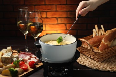 Photo of Woman dipping piece of broccoli into fondue pot with melted cheese at wooden table with wine and snacks, closeup