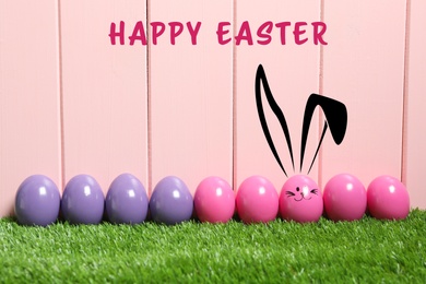 One egg with drawn face and ears as Easter bunny among others on green grass against pink wooden background