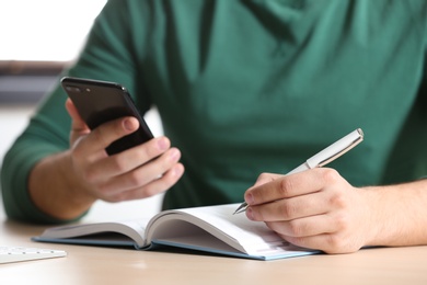Young man with smartphone writing in notebook at table, closeup