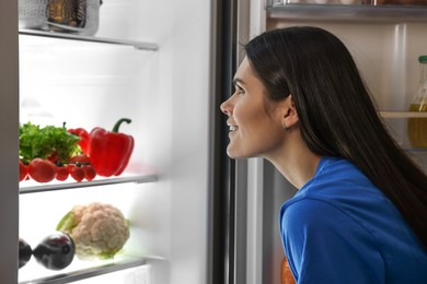 Photo of Young smiling woman looking into modern refrigerator at night